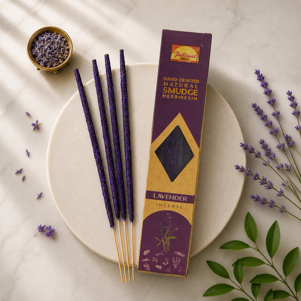 Lavender incense sticks in a purple and gold box labeled Parimal's Hand Crafted Natural Smudge Herbs' on a white marble plate, styled with dried lavender, green leaves, and a small bowl of lavender buds.