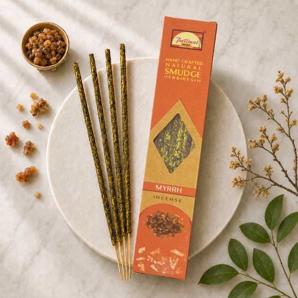 A styled flat-lay of a box of Parimal Myrrh Incense sticks on a white marble surface. The orange and gold packaging stands next to four incense sticks with green glitter, which are placed on a white ceramic plate.