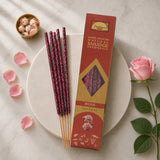 A styled flat-lay of Patrimoni Rose incense sticks and their packaging box on a round white marble plate, surrounded by pink rose petals, a pink rose, and a small gold bowl of rose buds on a light marble background.