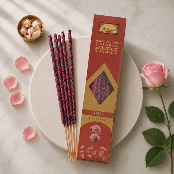 A styled flat-lay of Patrimoni Rose incense sticks and their packaging box on a round white marble plate, surrounded by pink rose petals, a pink rose, and a small gold bowl of rose buds on a light marble background.
